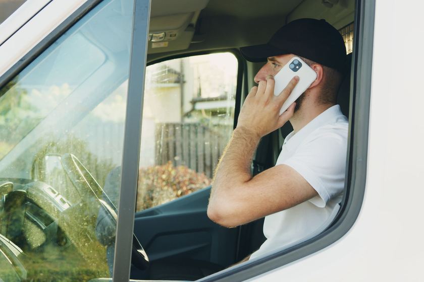 A tradesman in a cap and white polo sits in the driver’s seat of his white work van, taking a call on his mobile between jobs