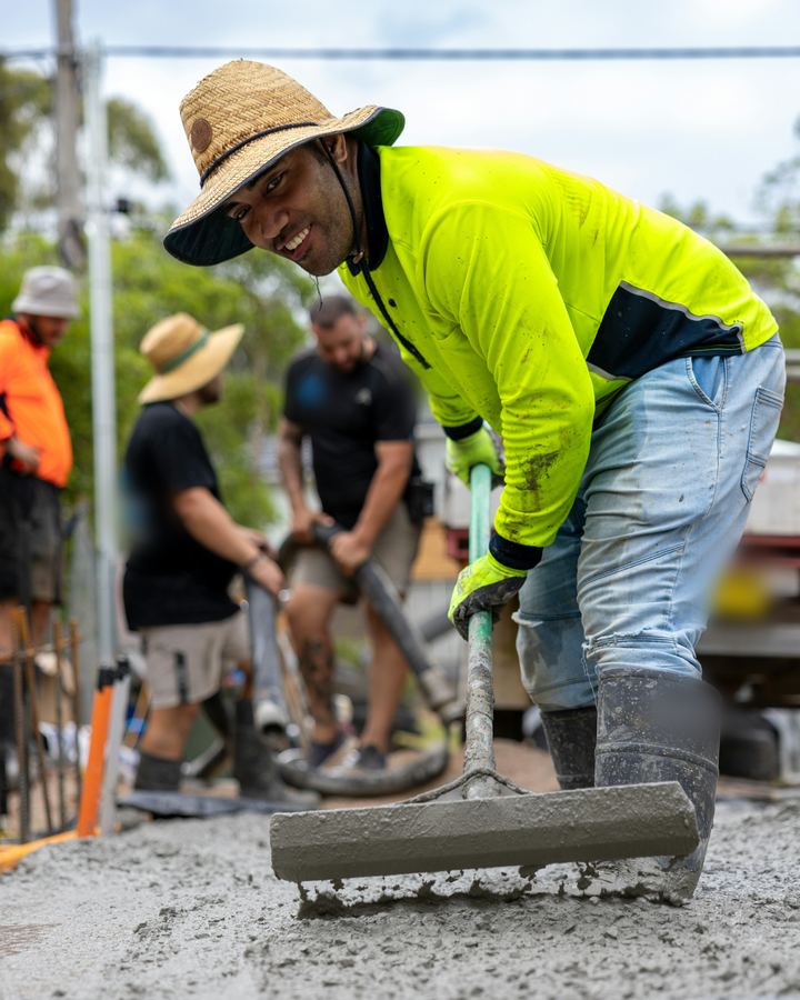 A smiling concreter in a broad-brim straw hat and yellow hi-vis shirt screeds a fresh concrete slab while his crew works the pump hose behind him